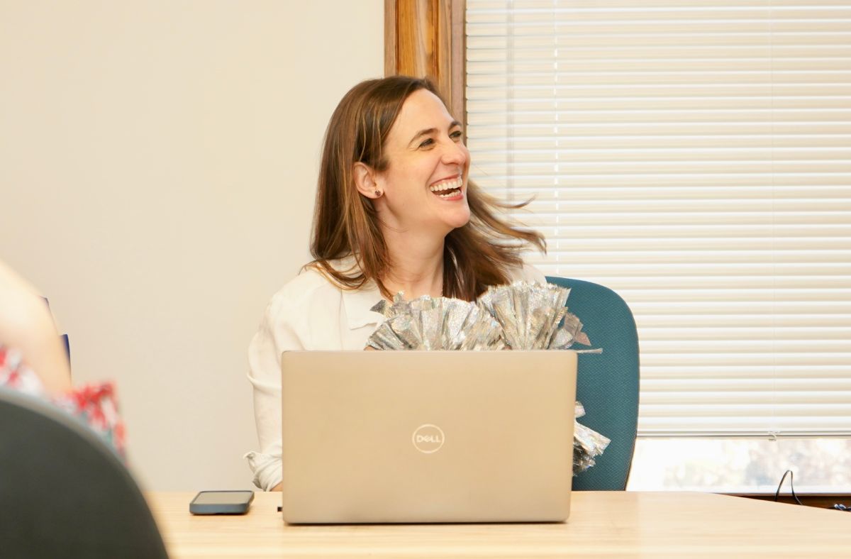 Photo of woman smiling in a team meeting at the Omlie house shaking pom poms with her computer in front of her.